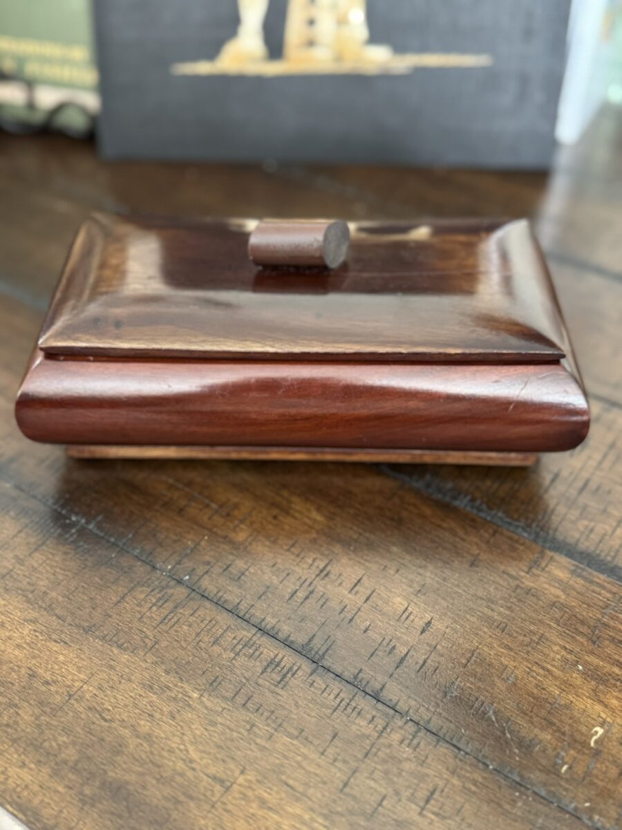 A small, rectangular wooden box with a sloped lid and a round knob sits on a wooden table. The box has a polished, dark brown finish, and blurred objects can be seen in the background.