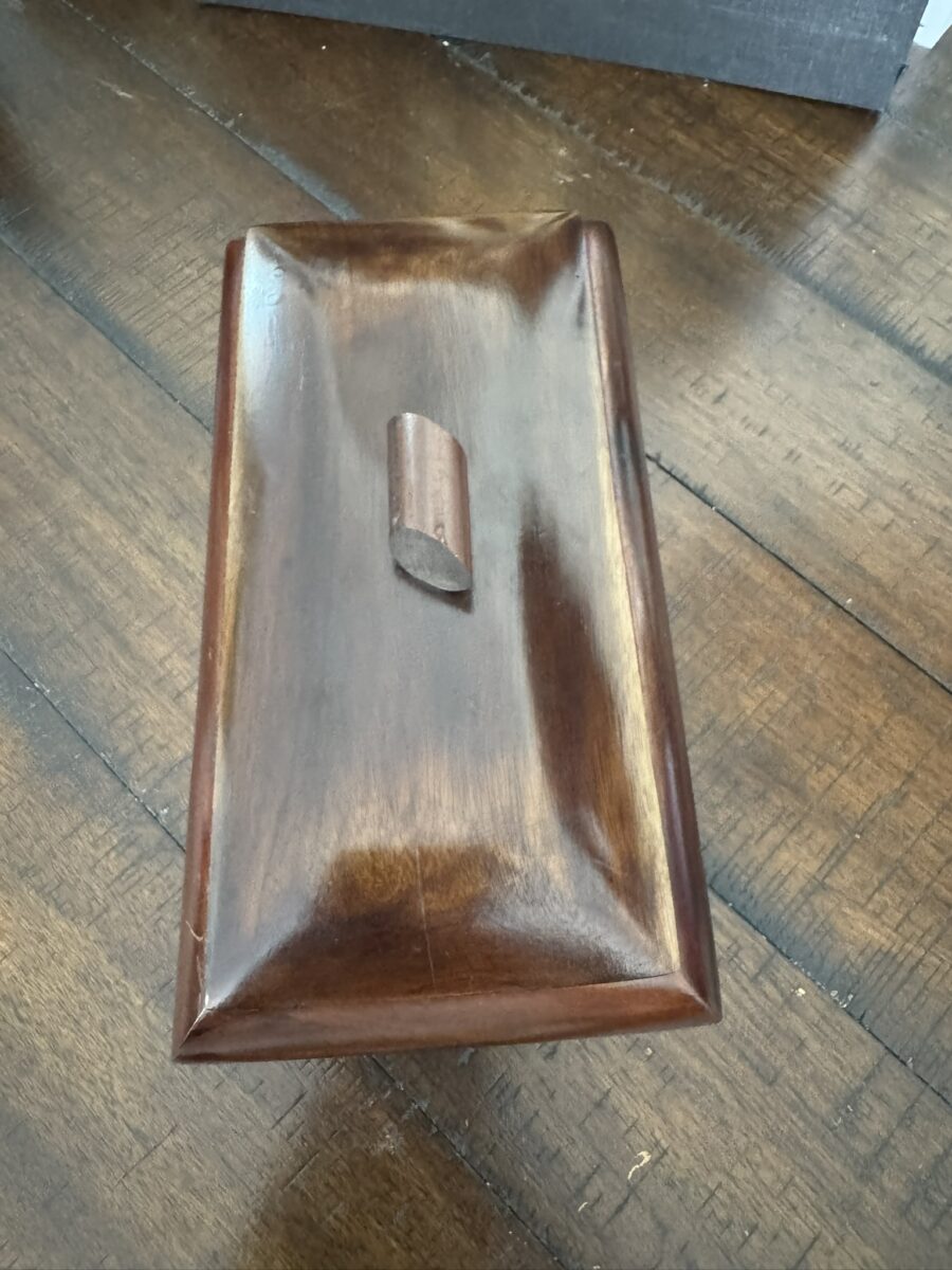 A rectangular wooden box with a glossy finish sits on a wooden floor. The box has a domed lid with softened edges and a small, upright, cylindrical handle in the center, angled slightly to the left. The wood grain is visible and polished.