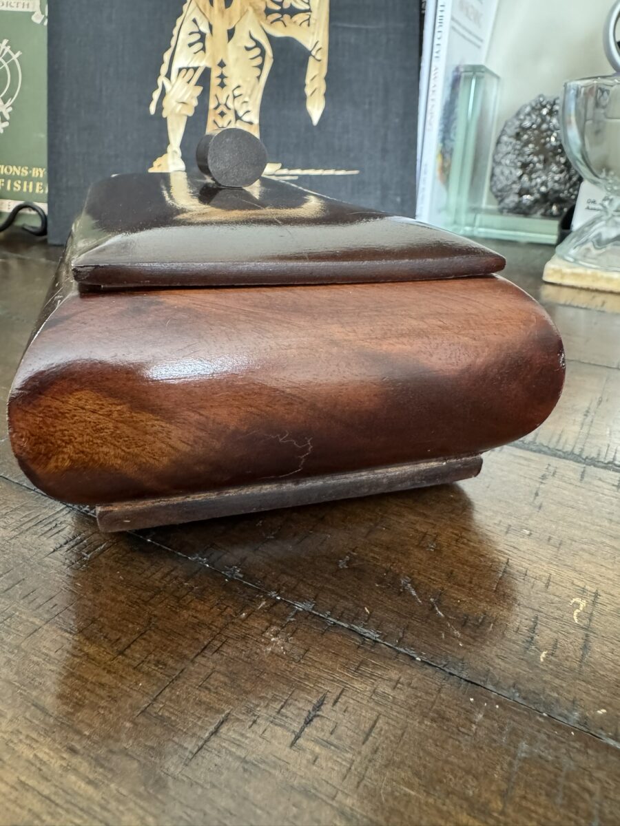 A close-up of a polished wooden box with rounded edges, resting on a dark wood surface. The lid is slightly raised, revealing a metal knob on top. Background items include books, a crystal, and various decorative objects.