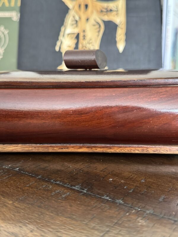 A close-up of a polished wooden box with a curved lid and a cylindrical handle on top, sitting on a wooden surface. In the background, partially visible, are books, one with a gold illustration on a black cover.
