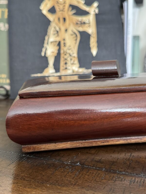 Close-up of a polished wooden box with a hinged lid, resting on a wooden surface. In the blurred background, there is a gold-colored decorative figure and partially visible books or objects. The image focuses on the wood grain and smooth finish of the box.