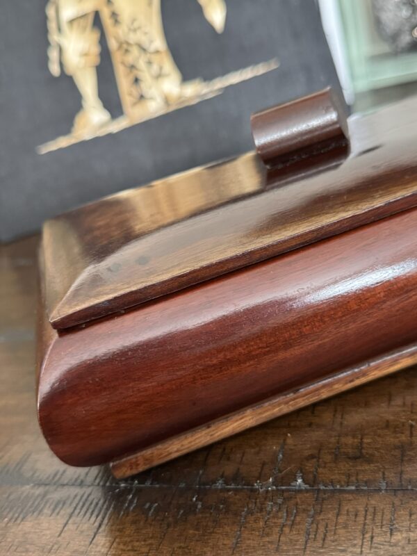 A close-up of a polished wooden box with a hinged lid, sitting on a dark wood surface. In the background, a blurred decorative panel features a gold figure. The image highlights the box’s glossy finish and smooth, rounded edges.