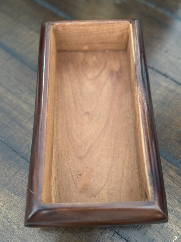 A close-up of an empty rectangular wooden box with smooth, polished edges and a natural wood grain. The box is placed on a wooden surface with a visible linear texture. The lighting highlights the warm tones of the wood.