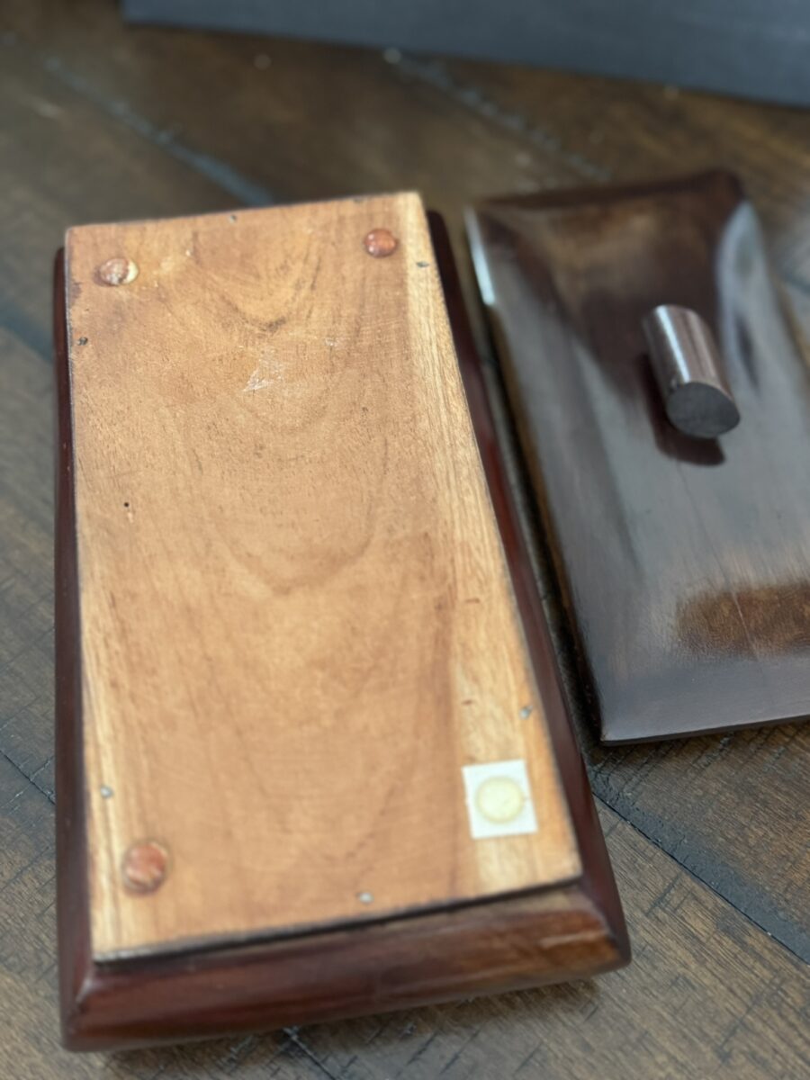 A rectangular wooden box with its lid removed sits on a brown wood surface. The box’s base shows screws and two small felt pads in opposite corners. The lid, with a smooth dark brown knob, rests nearby, revealing the lighter wood inside the box.
