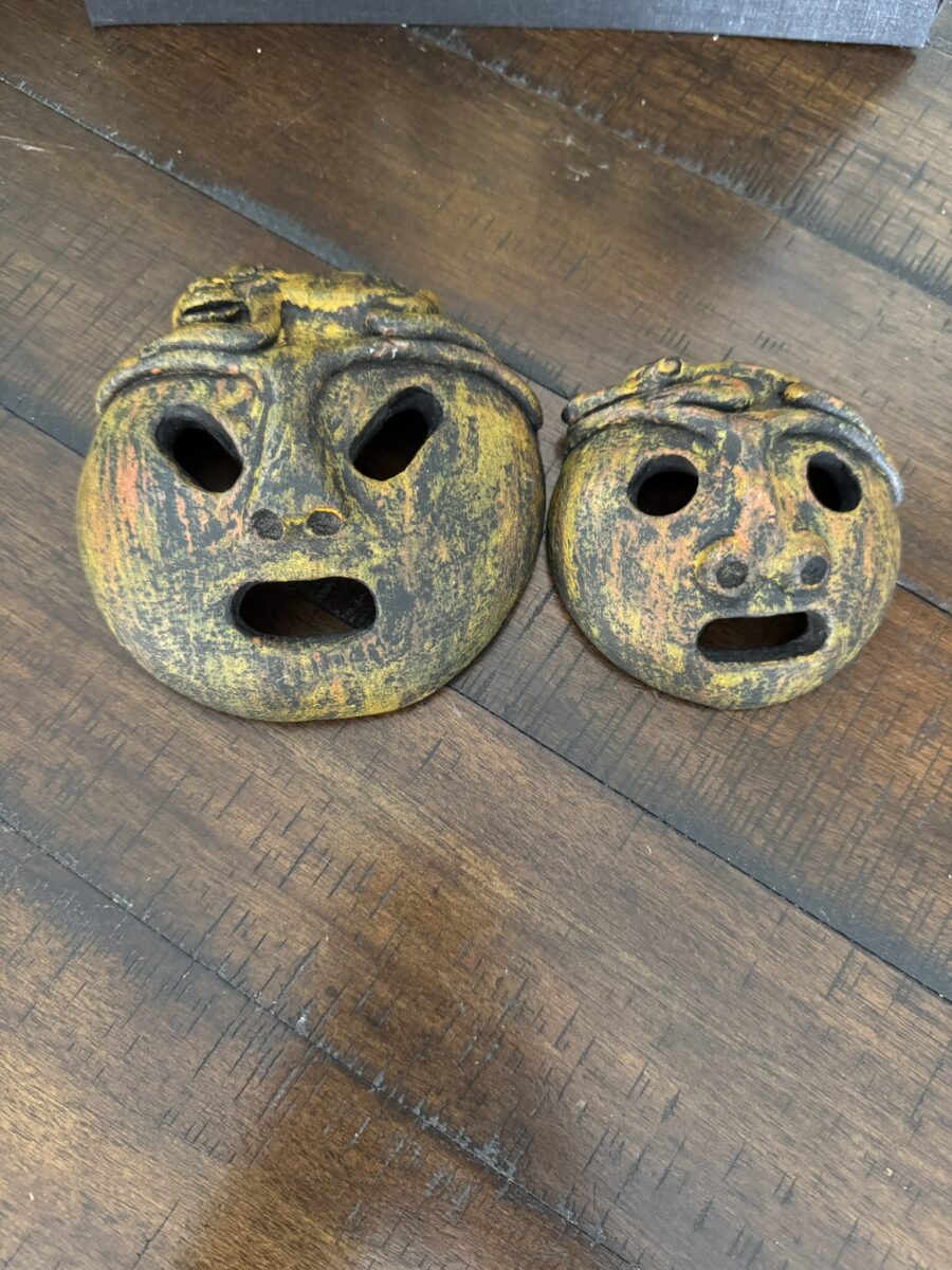 Two round, rustic, and weathered masks with distressed yellow and orange paint rest on a wooden surface. Both masks have hollow eyes and open mouths expressing shock or surprise, with small decorative elements on their foreheads.