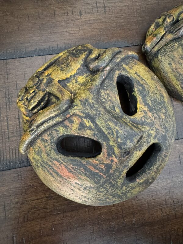 A round, textured stone mask with an open mouth and eye holes sits on a wooden surface. The mask has earthy yellow and orange hues and features a sculpted frog resting on its forehead, blending naturally into the mask’s surface.