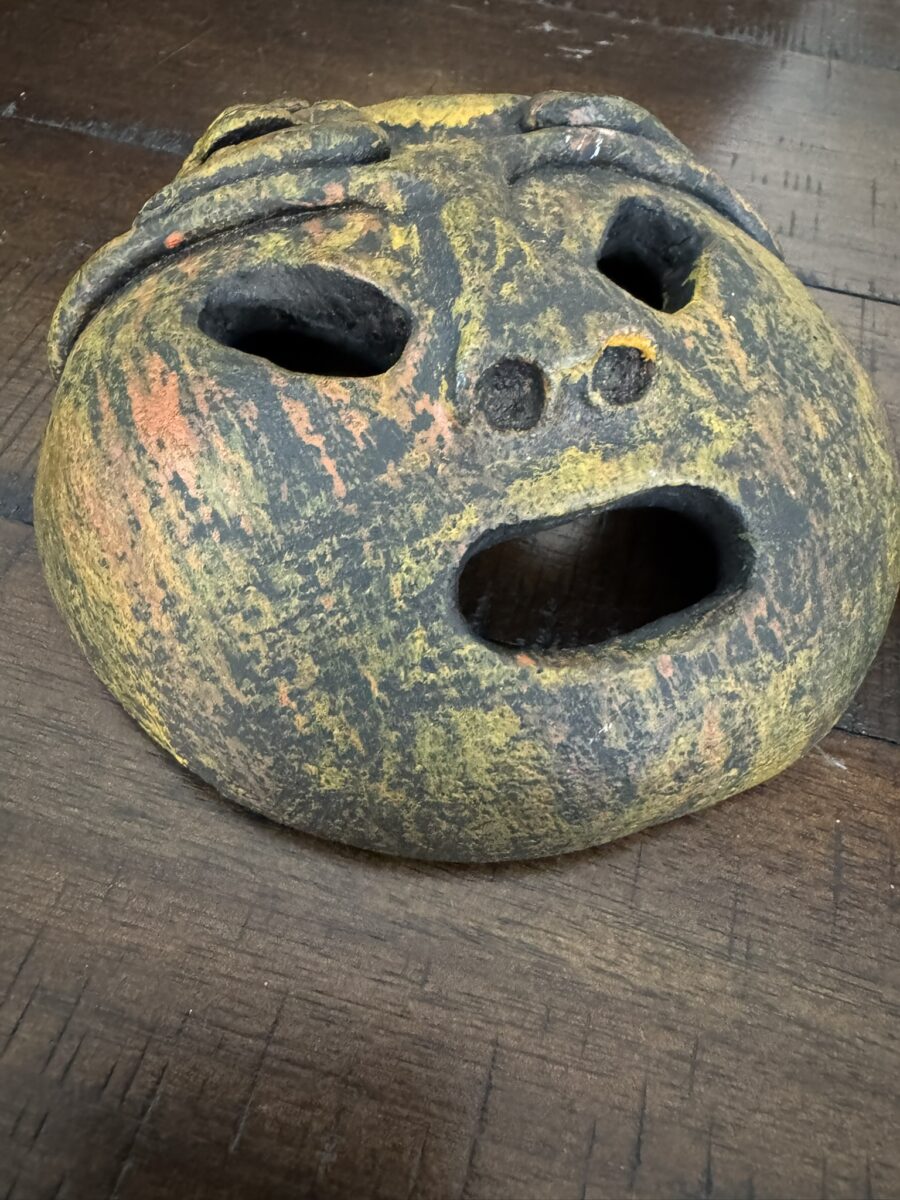 A weathered, round, yellowish-brown clay mask with a wide open mouth, two eye holes, and two small holes for the nose rests on a wooden surface. The mask has a rough, textured surface with faint orange and black patches.