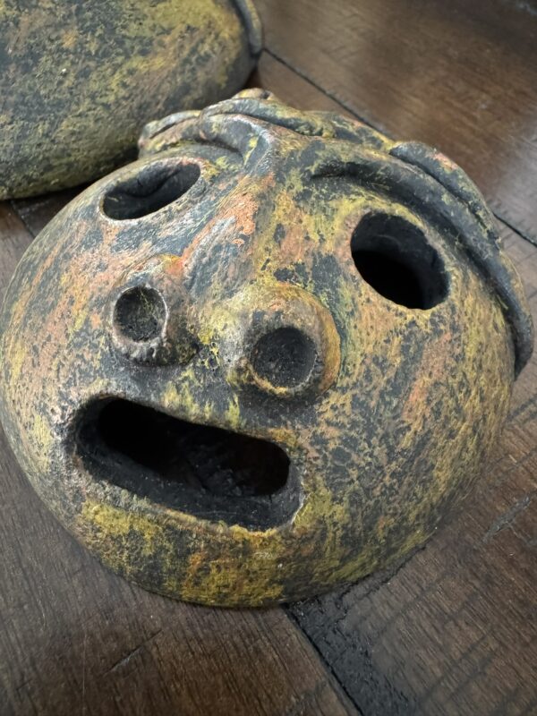 A round, weathered ceramic mask with a wide open mouth, large round nose, and hollow eyes. The surface is mottled with yellow, orange, and brown tones. The mask rests on a dark wooden surface, with part of another similar object visible behind it.