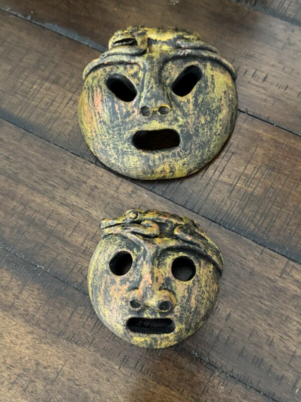 Two round, rustic masks with open mouths and eyes, painted with mottled yellow, orange, and black. Both have a snake motif across their brows. The masks rest on a dark wooden surface. The top mask is larger; the bottom one is smaller.
