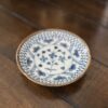 A small, round ceramic plate with intricate blue floral and geometric patterns sits on a wooden surface. The plate has a slightly raised rim and features a central floral motif surrounded by detailed designs. The overall style appears to be inspired by traditional pottery.