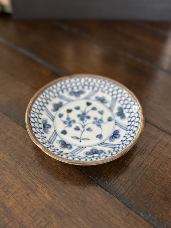 A small, round ceramic plate with intricate blue floral and geometric patterns sits on a wooden surface. The plate has a slightly raised rim and features a central floral motif surrounded by detailed designs. The overall style appears to be inspired by traditional pottery.