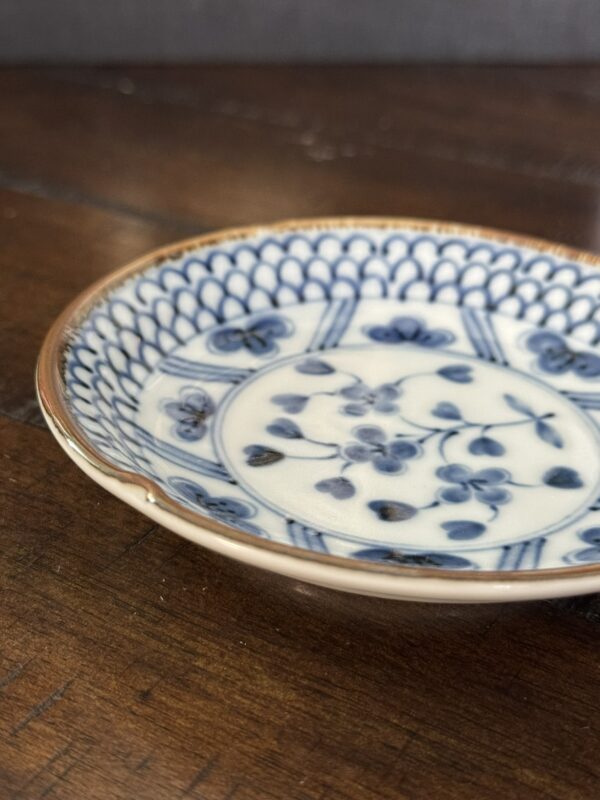 A close-up of a small, shallow porcelain dish with a blue floral and geometric pattern on a white background. The rim is lightly brown, and one section shows a visible chip. The dish rests on a dark wooden surface.