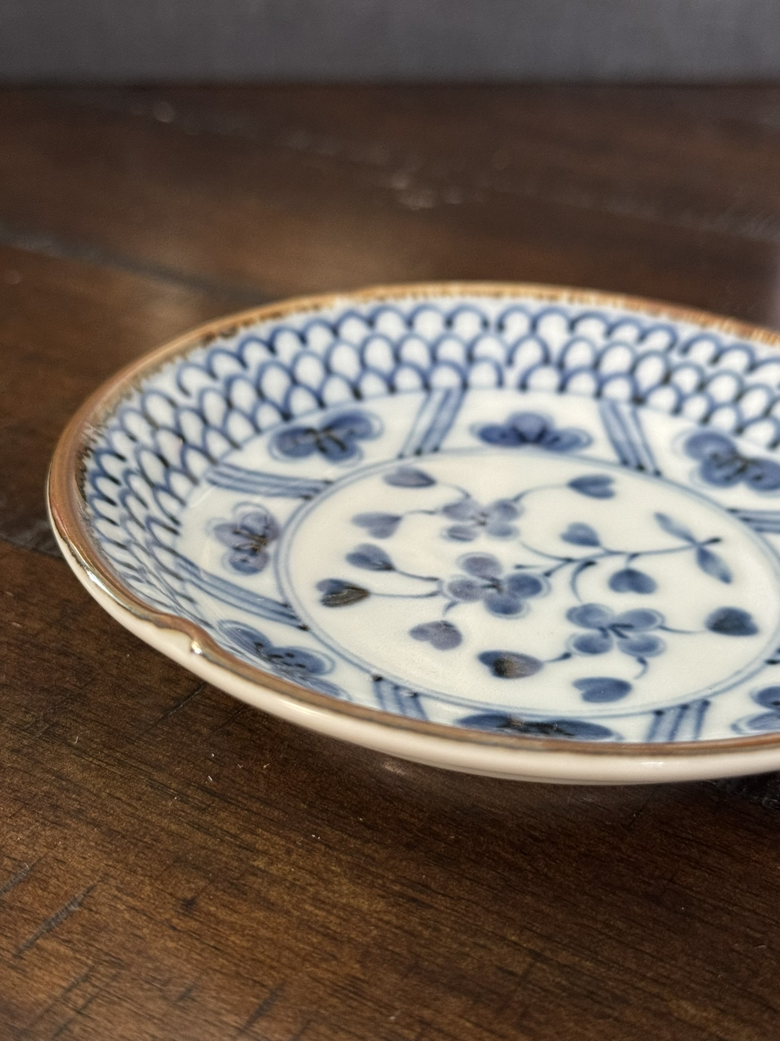 A close-up of a small, shallow porcelain dish with a blue floral and geometric pattern on a white background. The rim is lightly brown, and one section shows a visible chip. The dish rests on a dark wooden surface.