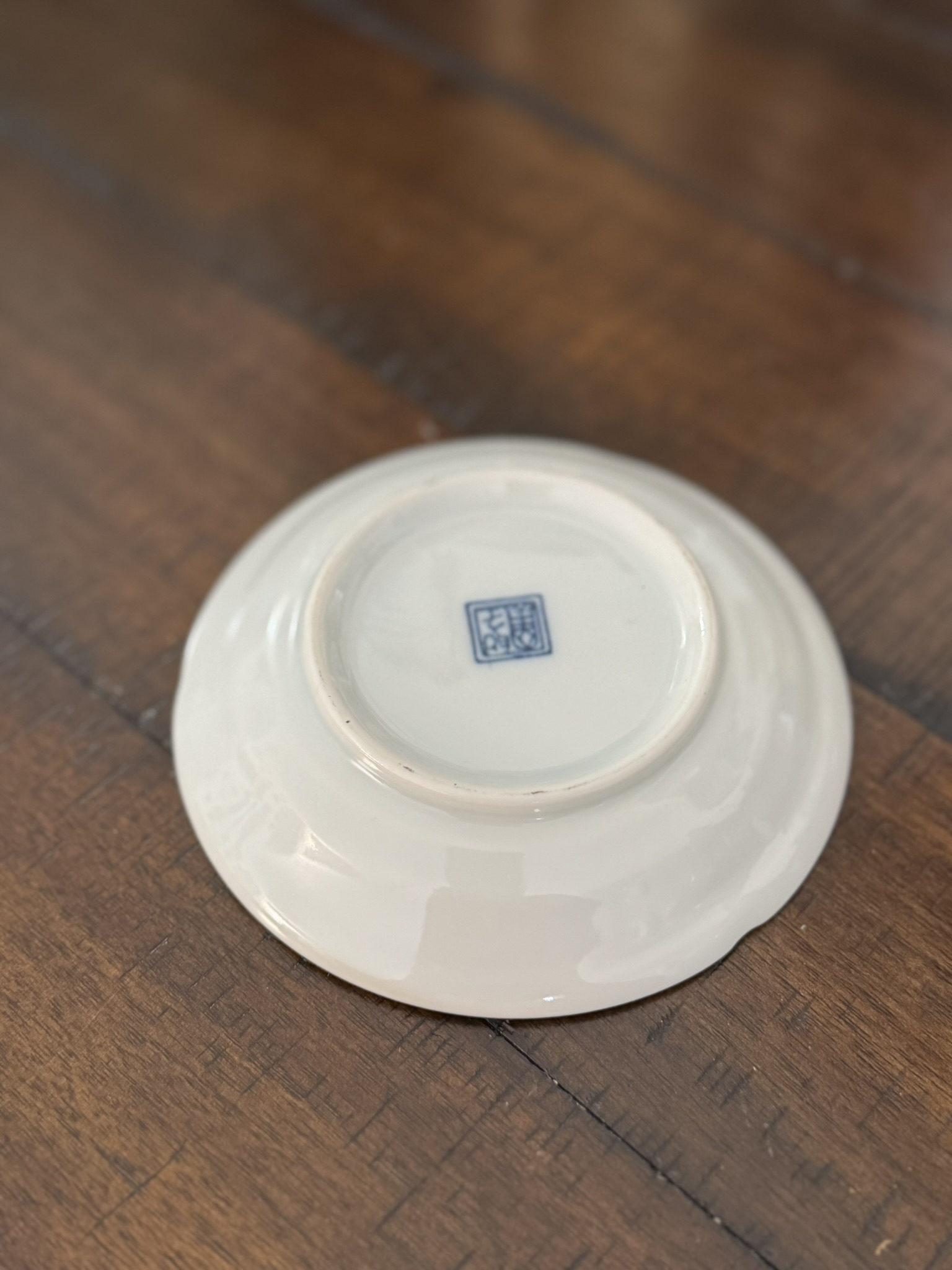 A small, white ceramic bowl is shown upside down on a wooden surface. The base features a blue square seal with Chinese characters. The bowl has a smooth, glossy finish and a slight chip on the rim. The wooden surface is polished and brown.
