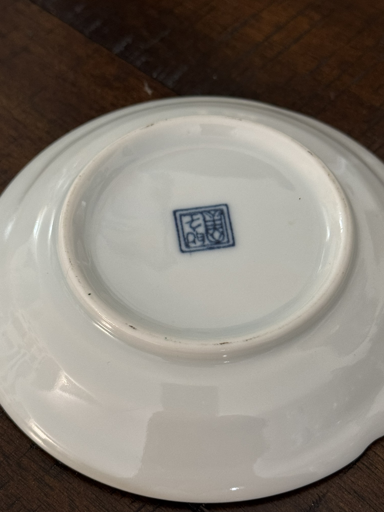 A white porcelain dish is shown upside down on a wooden surface. At the center of its base, there is a small, blue square stamp featuring Chinese characters. The edges of the dish are smooth, and the glaze is shiny.