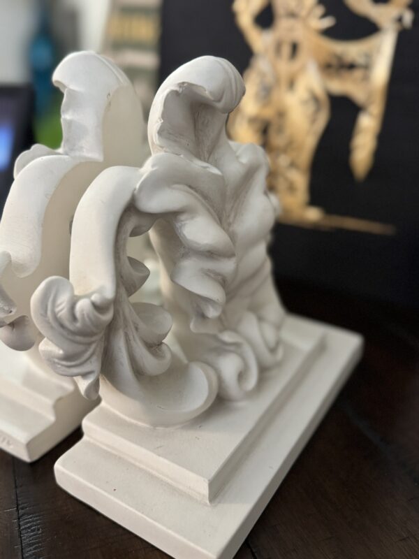 Two ornate, white plaster bookends with swirling leaf designs sit on a dark wooden surface. In the blurred background, there is a decorative gold figure on a black surface. The focus is on the intricate details of the bookends.