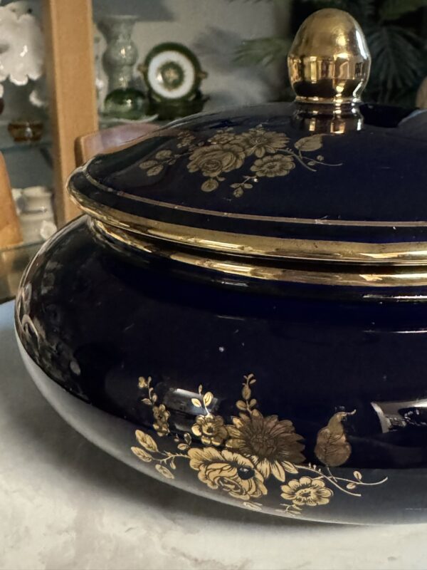 A glossy, dark blue ceramic tureen with a lid, decorated with gold floral patterns and a gold knob. The tureen sits on a light surface, with blurred background elements including glassware and wooden decor.