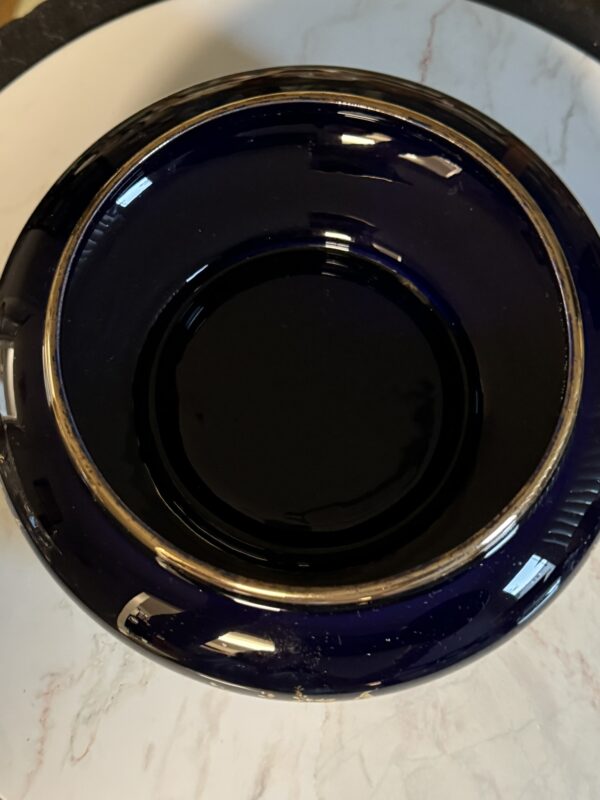 A deep, round, dark blue bowl with a gold rim sits upside down on a white marble surface. Reflections of light are visible on the glossy surface of the bowl, and the bowl’s base is centered in the image.