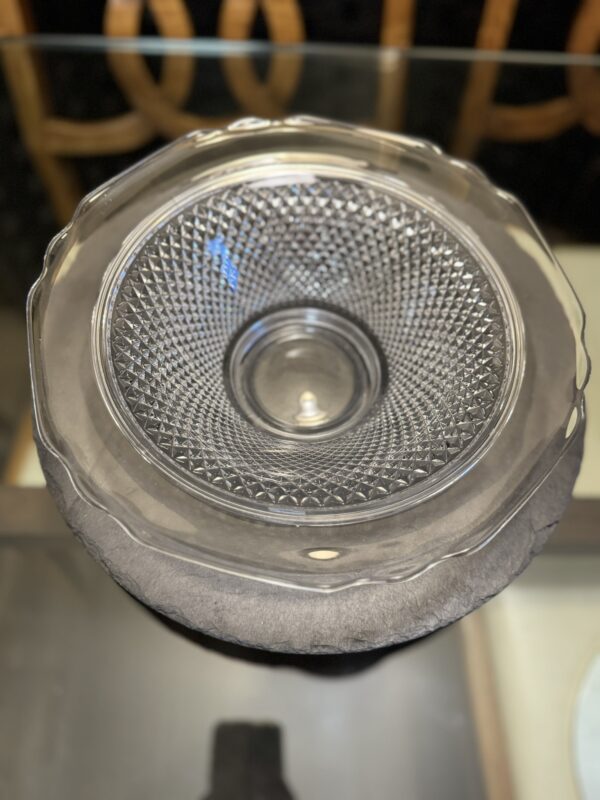A clear glass bowl with a textured, diamond-patterned interior sits atop a larger, smooth glass plate on a dark round surface. Both are centered in the image, with wooden chair backs and a reflective tabletop visible in the background.