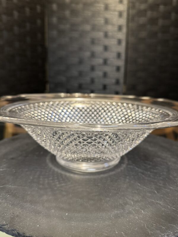 A clear glass bowl with a diamond-patterned texture sits on a dark, round surface. The bowl has a wide rim and a shallow depth, and the background features a black, woven design. Light reflects off the bowl, highlighting its intricate pattern.