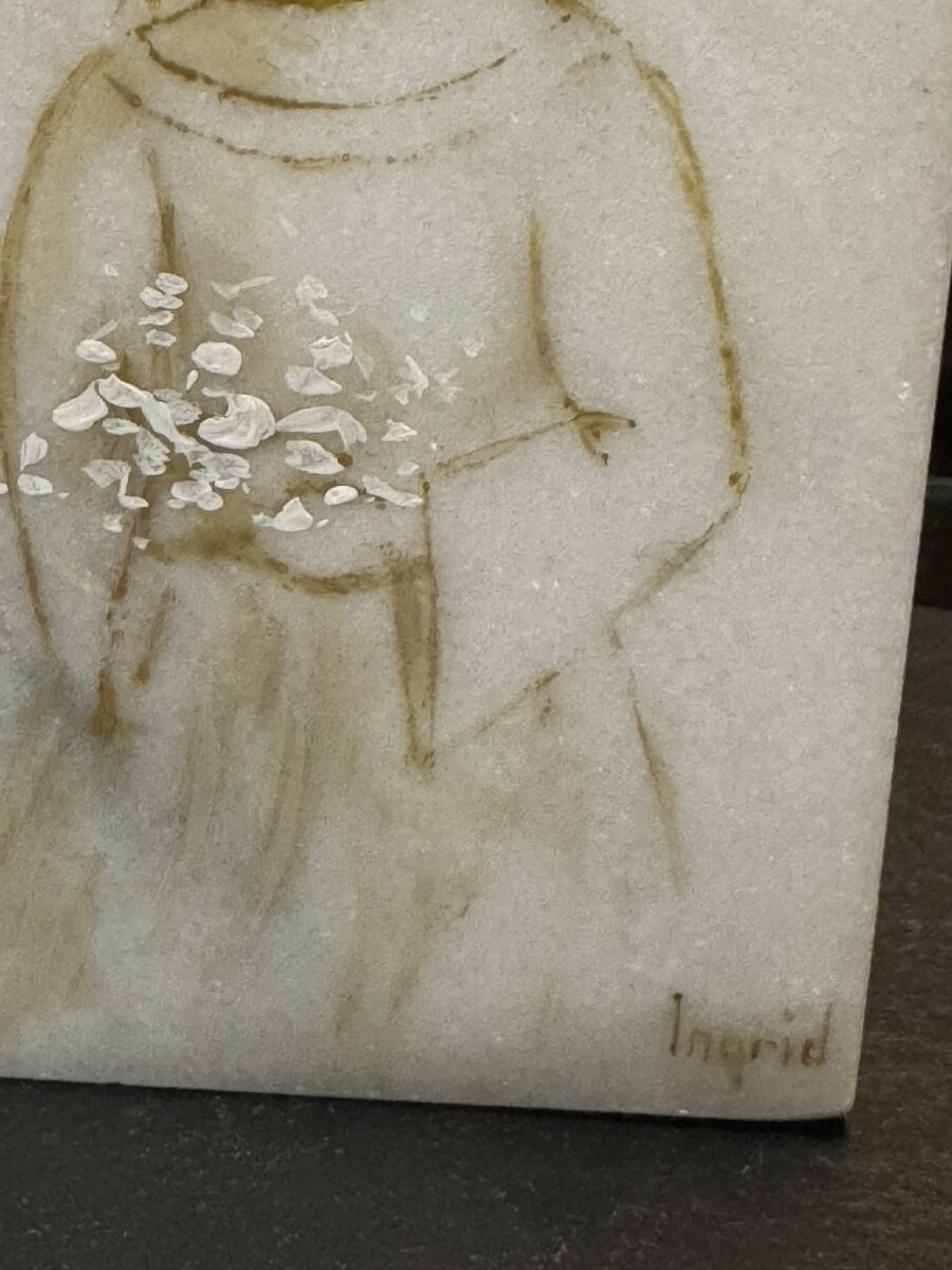 A simple line drawing on a stone surface depicts a figure in a robe holding white flowers. The figure’s face is not shown, and only the upper body is visible. The artist’s signature, Ingrid, appears in the bottom right corner.