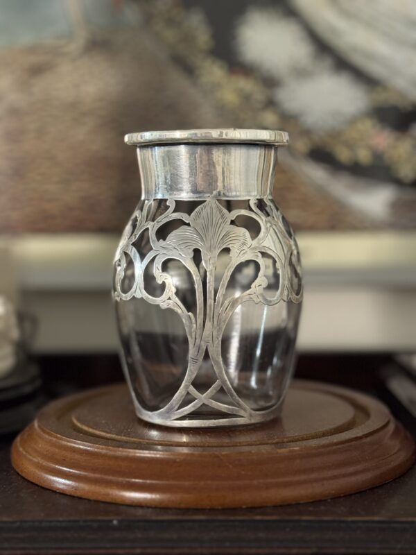 The Miniature Silver Overlay Vase showcases an Art Nouveau floral design in ornate silver on glass. It rests on a polished wooden base with a softly blurred background, emphasizing its detailed craftsmanship and decorative appeal.