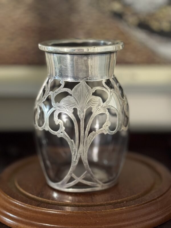 The Miniature Silver Overlay Vase, a clear vase with ornate silver Art Nouveau floral patterns, stands on a round polished wooden surface with a blurred painting in the background.