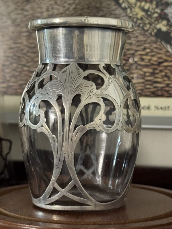 The Miniature Silver Overlay Vase features an Art Nouveau floral design with tall, stylized iris flowers and curving stems, displayed on a wooden surface with a blurred framed artwork in the background.