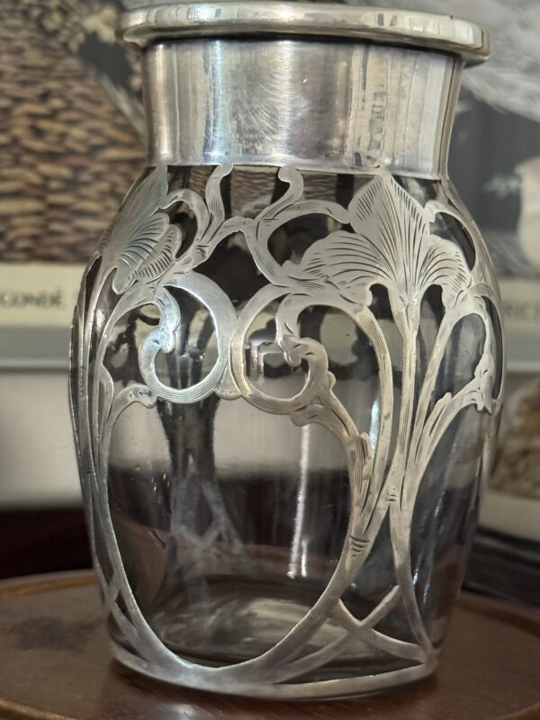 The Miniature Silver Overlay Vase features clear glass with a wide silver rim and intricate Art Nouveau floral silver overlay of flowing lines and stylized flowers. Shown on a wooden surface with a blurred art poster in the background.