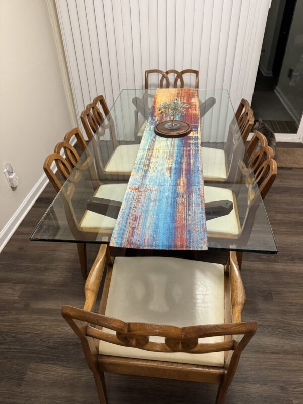 A glass dining table with six John Stuart chairs sits on a dark wood floor. A colorful, abstract-painted wooden runner in blue, orange, and yellow lies at the center, topped with a round metal tray. Vertical blinds complete the scene.