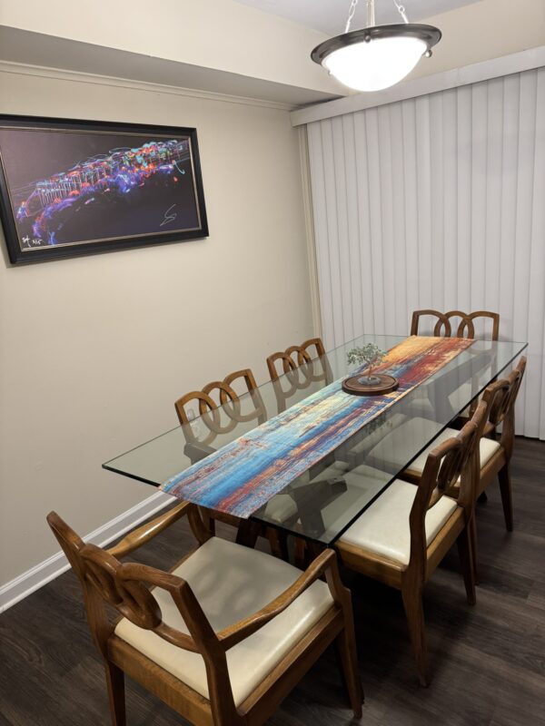 A dining room with a glass-top table and a set of 6 John Stuart Chairs with cream cushions, a colorful table runner, small plant centerpiece, light walls, vertical blinds, dark wood flooring, and a modern pendant light.