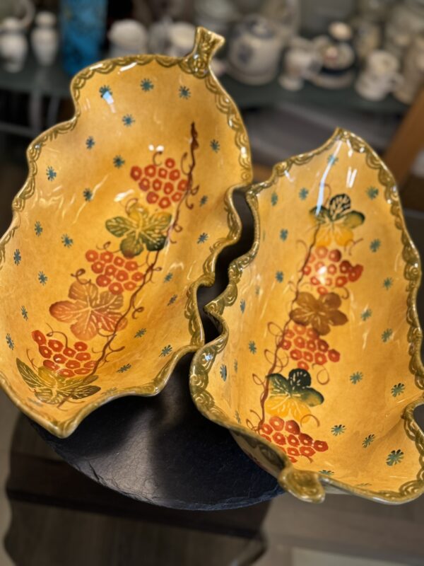 Two Italian Oblong MCM Serving Dishes, ceramic with scalloped edges and leaf shapes, display yellow bases decorated with red, green, and blue floral and berry patterns. They sit side by side on a dark surface; pottery is blurred in the background.