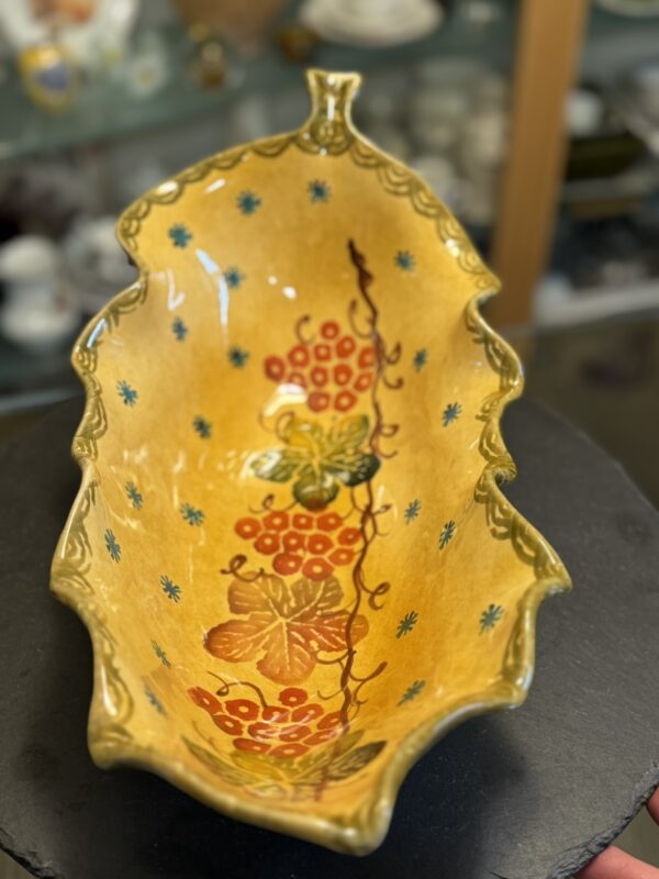 The Italian Oblong MCM Serving Dish is a yellow, leaf-shaped ceramic piece with scalloped edges and red, green, and blue floral vines. It rests on a black surface with blurred shelves of other ceramics in the background.