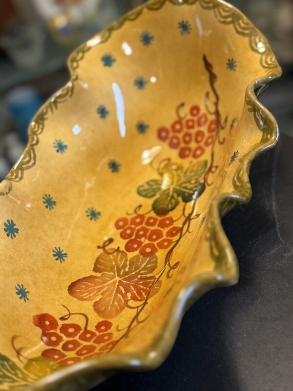 A close-up of an Italian Oblong MCM Serving Dish shows yellow, wavy edges with hand-painted red berries, green leaves, yellow butterflies, blue floral accents, and a green border. The vibrant dish rests on a dark surface.
