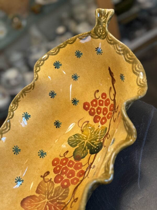 A close-up of the Italian Oblong MCM Serving Dish shows its scalloped rim, hand-painted red grapes, green vine leaves, and blue floral patterns on a warm yellow background, with a handle at one end.