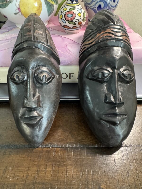 The African Style Mask Pair, featuring ornate headdresses and detailed elongated faces, sits side by side on a wooden table, with colorful ceramic vases, a pink book, and a partially titled “OF” book displayed behind them.