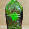 The Cure Bottle, a vintage green glass piece with embossed “Safe Kidney & Liver Cure” and “Rochester, N.Y.,” rests on a beige surface, displaying age-related wear and subtle iridescence.