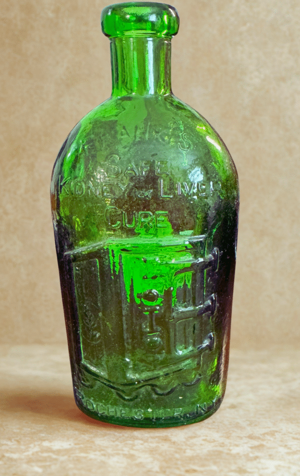The Cure Bottle, a vintage green glass piece with embossed “Safe Kidney & Liver Cure” and “Rochester, N.Y.,” rests on a beige surface, displaying age-related wear and subtle iridescence.