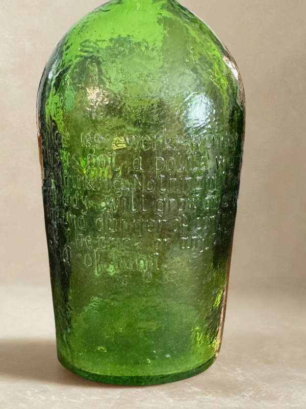 A green, textured glass Cure Bottle stands upright against a plain light beige background. Its uneven, bubbly surface features embossed text that's hard to read due to the texture and lighting, giving the bottle an antique appearance.