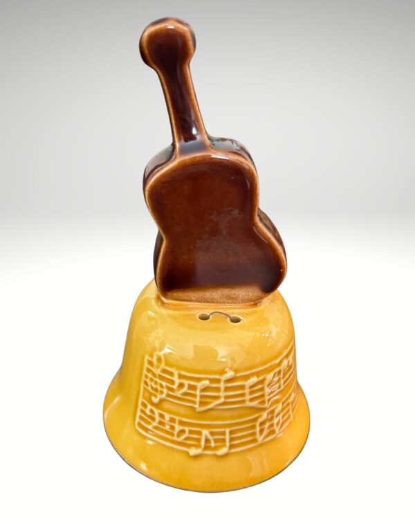 The Opryland Guitar Bell is a yellow ceramic bell with a brown guitar-shaped handle, inspired by Opryland. Embossed musical notes decorate its surface, making it a unique collectible. The background is a plain, light gradient.