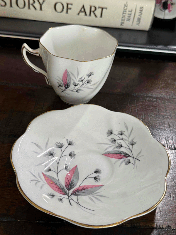The Vintage Teacup Set, with a white cup and saucer trimmed in gold and a black, gray, and pink leaf pattern, rests on a dark wooden table. Behind it are two large books: HISTORY OF ART and PRENTICE HALL ARTWAYS.