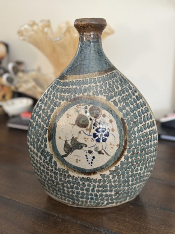 A blue and tan Tonala Vase with a rounded body, narrow neck, dotted texture, and central circular panel featuring hand-painted floral and berry designs in earthy tones and blue sits on a dark wooden table.