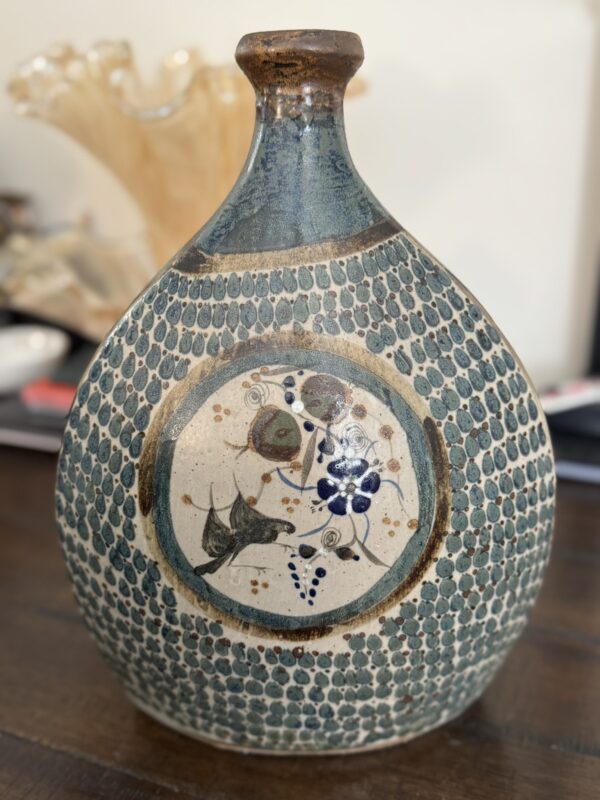 The Tonala Vase is a round, blue-green ceramic piece with textured dotted patterns and a central circular design featuring painted birds, berries, and foliage in earthy tones and blue. It’s photographed against a softly blurred indoor background.