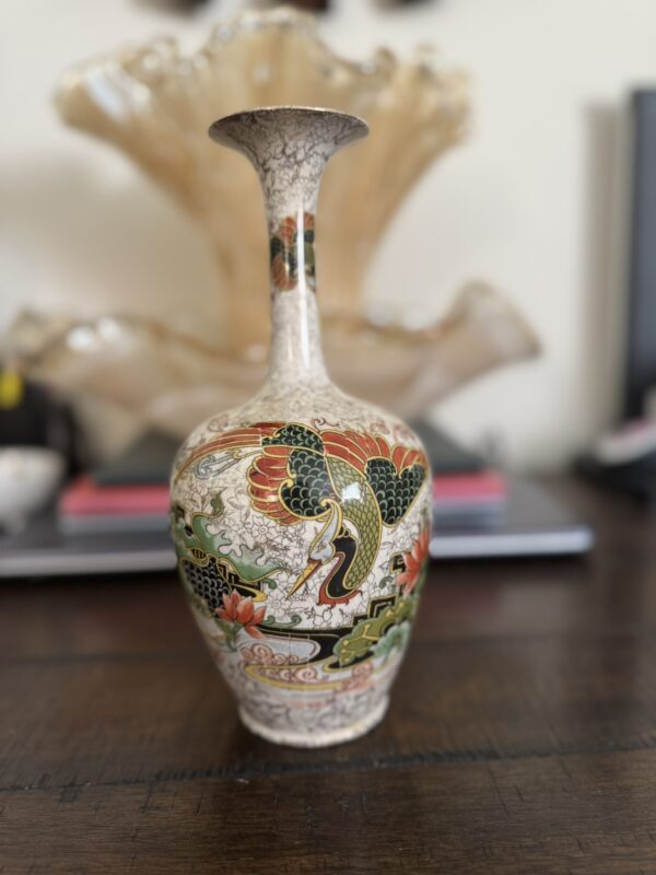 The Chinese Blue & White Miniature Vase features vibrant painted designs, including a colorful bird with spread wings, flowers, and gold accents. It is displayed on a dark wooden table with blurred decor and books in the background.