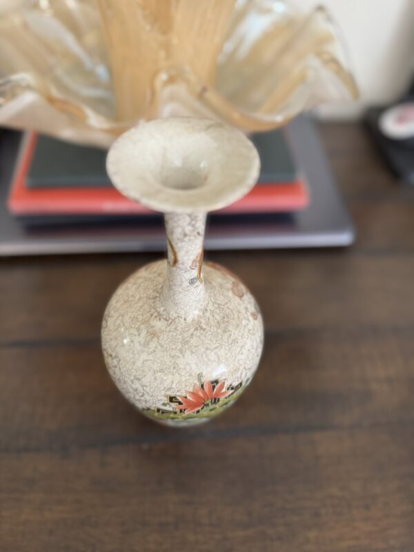 A narrow-necked ceramic vase with a crackled beige glaze and red floral design sits on a wooden surface. Behind it are stacked books, a beige wavy-edged glass bowl, and the Chinese Blue & White Miniature Vase.