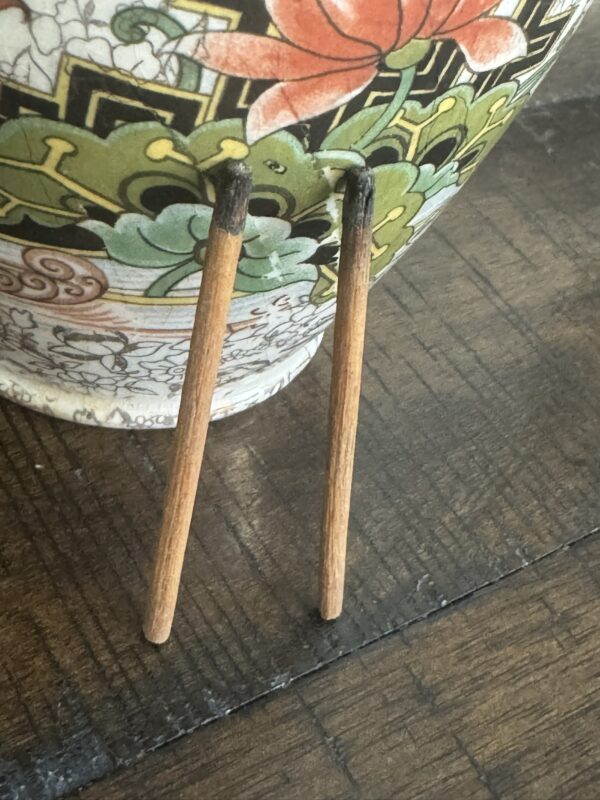 Two partially burned incense sticks with blackened tips lean in a V shape against a Chinese Blue & White Miniature Vase with colorful floral and green patterns, all resting on a wooden surface.