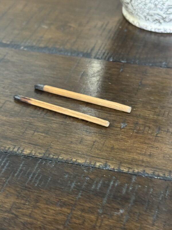 Two used matchsticks with burnt tips lie parallel on a warm, brown wooden surface. In the upper right corner, part of a white cup with a floral pattern—reminiscent of the Chinese Blue & White Miniature Vase—is visible. The wood shows visible grain and seams.