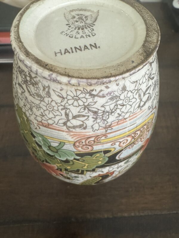 A Chinese Blue & White Miniature Vase rests upside down on a wooden surface, revealing a base stamped with ENGLAND, a crown emblem, and HAINAN. Its intricate blue floral patterns evoke the style of classic Chinese porcelain.