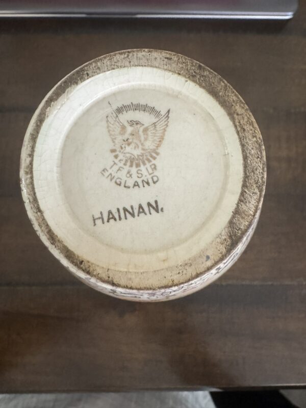 The Chinese Blue & White Miniature Vase features a ceramic base with a brown, aged rim, a maker’s mark showing a lion and “TJ&J S.L.R ENGLAND,” and “HAINAN.” printed beneath. The vase sits on a wooden surface.