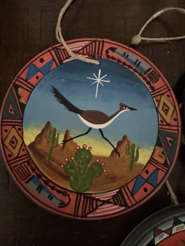 The Chinese Blue & White Miniature Vase features a desert scene with a running roadrunner, cacti, and rocks. Its border showcases geometric Southwestern patterns inspired by classic blue and white designs. A twine loop tops it; a white star shines above.